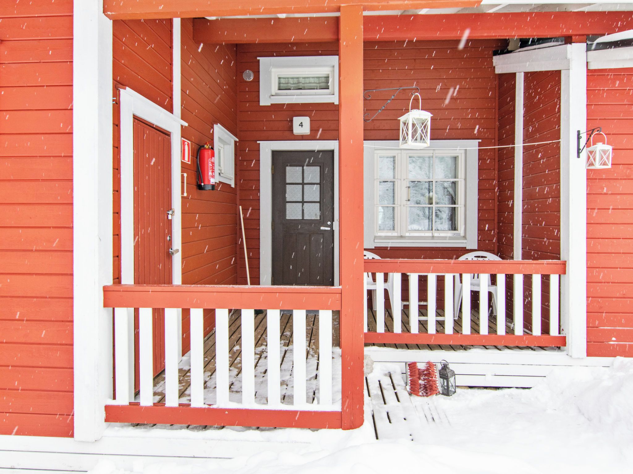 A red wooden house with a white porch, a door, and a window. Snow is falling.