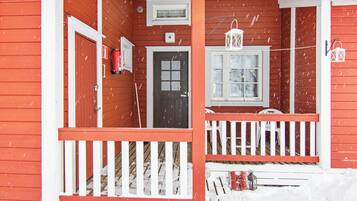 A red wooden house with a white porch, a door, and a window. Snow is falling.