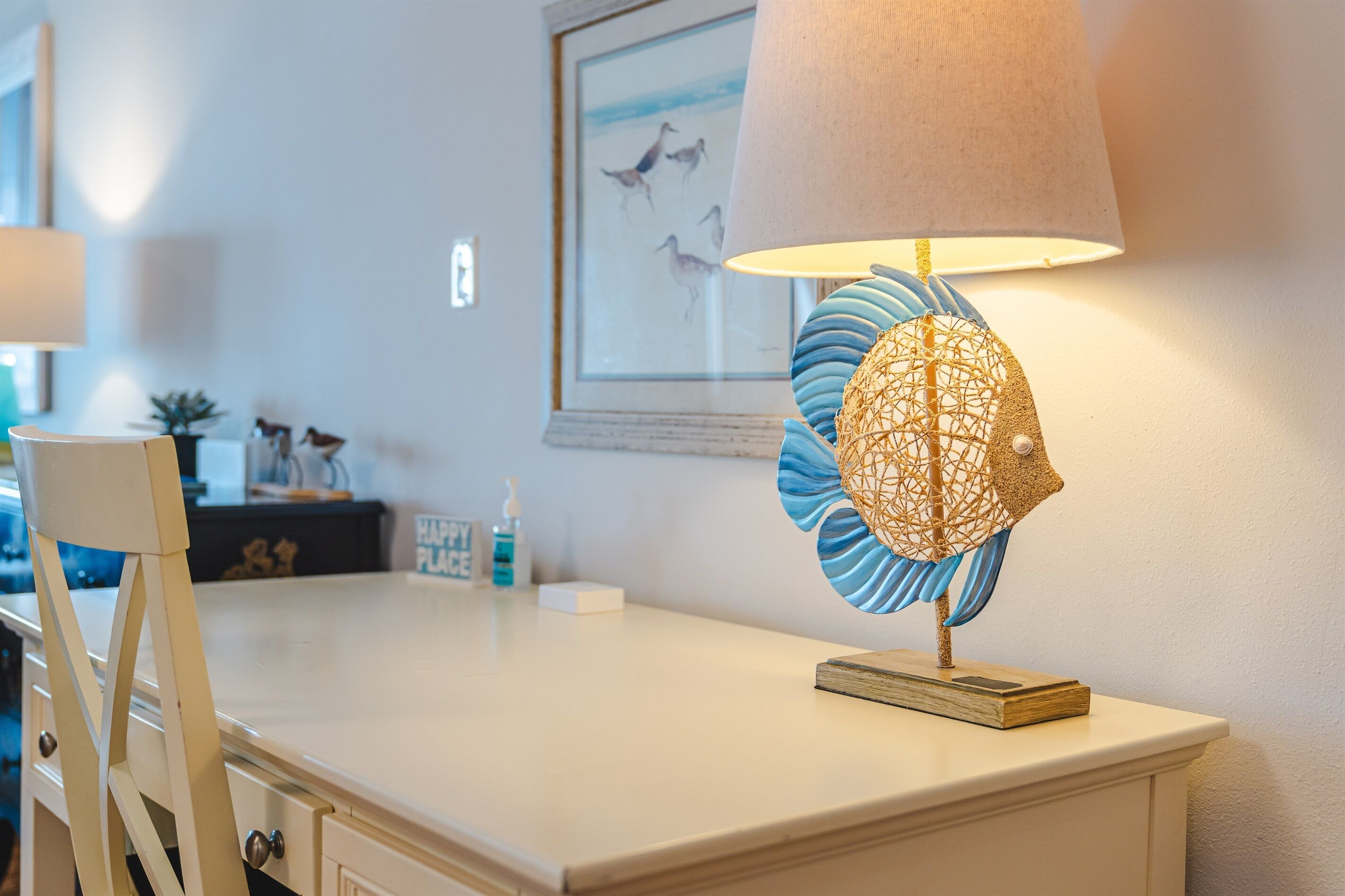 Een kamer met een wit bureau, een decoratieve lamp met blauw en goud design, een lijst met een foto aan de muur, en een klein plantje op het bureau.