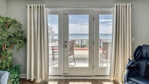A room with a sliding glass door leading to a balcony with a view of the sea. There is a potted plant and a leather sofa inside the room.