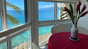 A room with a red table, a white vase with red flowers, and a view of the ocean through large windows.