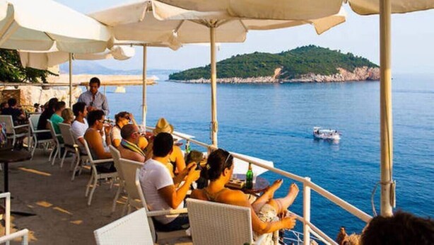 A seaside restaurant with outdoor seating overlooking the sea, a boat in the water, and a hilly landscape in the distance. - Apartment in center Old town (Dubrovnik)