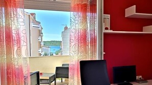 Ett rum med utsikt över byggnader och en balkong. Det finns ett skrivbord med en stol, en tv och en röd matta.