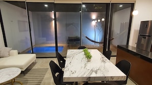 A modern living room with a marble table, black chairs, a white sofa, and a glass wall overlooking a pool area. - Casa BJ37 Mérida Norte, la mejor ubicación. (Mérida)