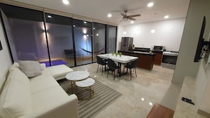 A modern living room with a sofa, a dining table with chairs, a kitchen area with appliances, and a large sliding glass door leading to a balcony. - Casa BJ37 Mérida Norte, la mejor ubicación. (Mérida)