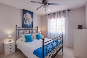 A bedroom with a bed, a nightstand, a ceiling fan, a window with curtains, and a painting on the wall.