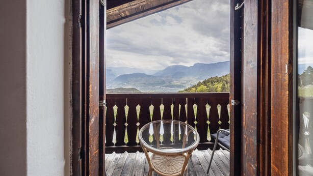 An open wooden door leading to a balcony with a wicker chair and a glass table, overlooking a mountainous landscape. - Apartment "Malgorerhof Lavendel" with Mountain View, Wi-Fi, Balcony & Shared Garden (Jenesien)