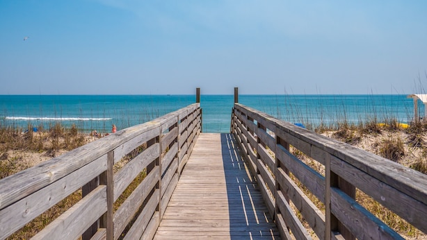 Ein hölzerner Steg führt zu einem Strand mit klarem, blauem Wasser und blauem Himmel.