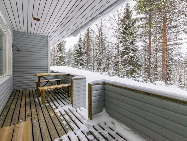 Una terraza de madera con un banco y una mesa, rodeada de árboles cubiertos de nieve.