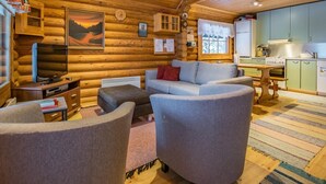 Ein gemütliches Blockhaus-Interieur mit einer Couch, Sesseln, einem Esstisch, einer Küchenzeile und einem Bild an der Wand.