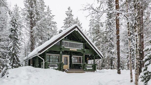 Eine Blockhütte mit Veranda und Schornstein, umgeben von schneebedeckten Bäumen.