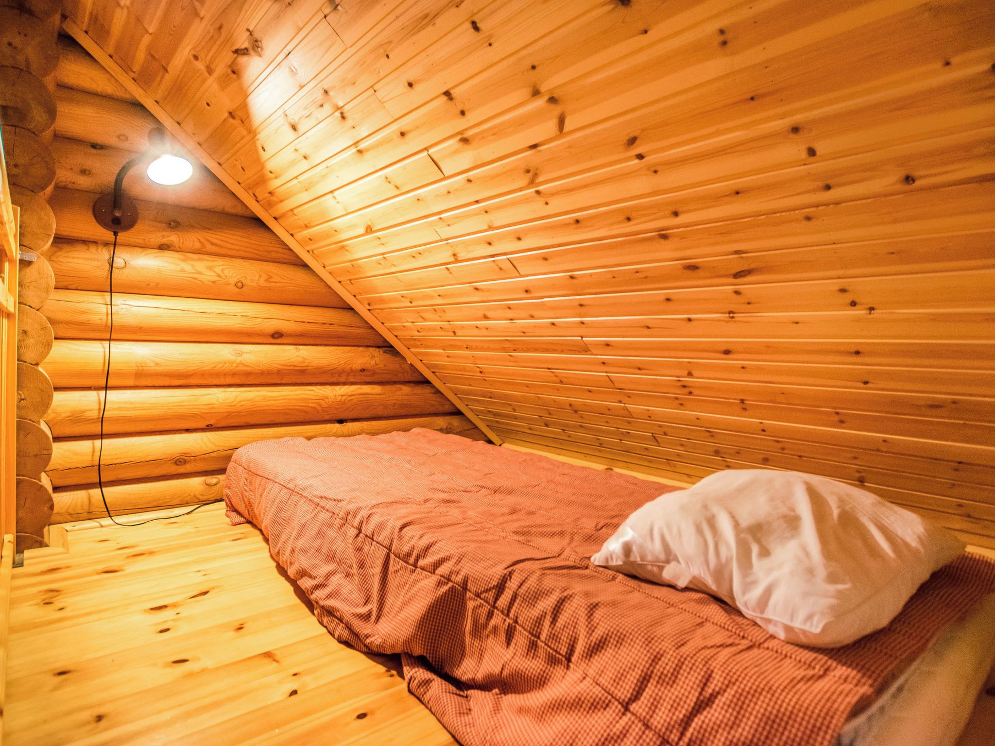 A log cabin room with a sloped ceiling, a single bed with a red blanket and white pillow, and a light fixture on the ceiling.