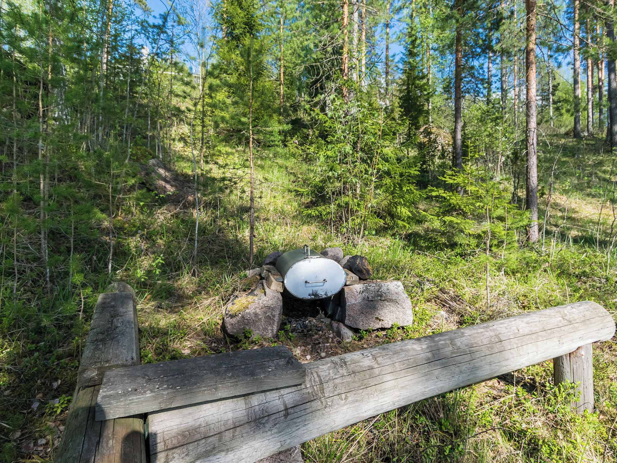 A forested area with a metal barrel and rocks, a wooden fence, and a clear sky.