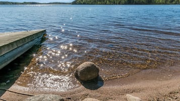 A wooden dock extending into a calm lake with clear water reflecting sunlight, surrounded by rocks and a forested shoreline.