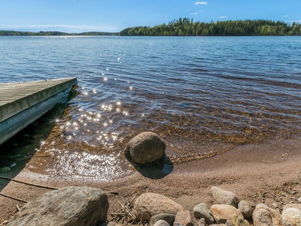 A wooden dock extending into a calm lake with clear water reflecting sunlight, surrounded by rocks and a forested shoreline.