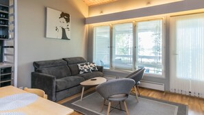 A modern living room with a grey sofa, a small round table, and a grey armchair. There are large windows with blinds, a wooden ceiling, and a painting on the wall. - Rantalinna 2 by Interhome (Ruokolahti)
