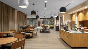A modern cafe with wooden tables and chairs, a central counter with a coffee machine, and a view of the city through the windows. - TownePlace Suites by Marriott Orlando Airport (Orlando)