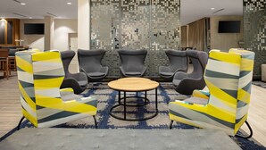 A modern lounge area with a round wooden table surrounded by grey and yellow striped armchairs, a patterned wall divider, and a flat-screen TV mounted on the wall. - TownePlace Suites by Marriott Orlando Airport (Orlando)