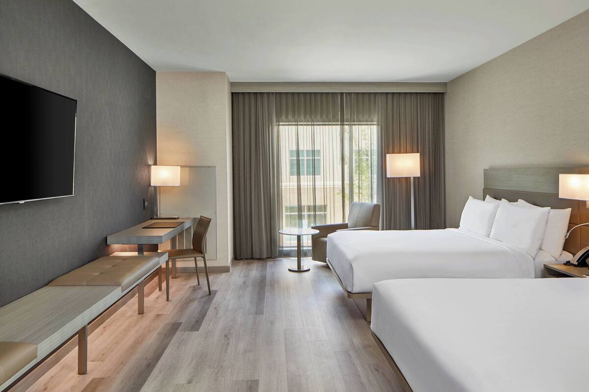 A hotel room with a TV, two beds, a desk, a chair, a table, and a window with curtains.