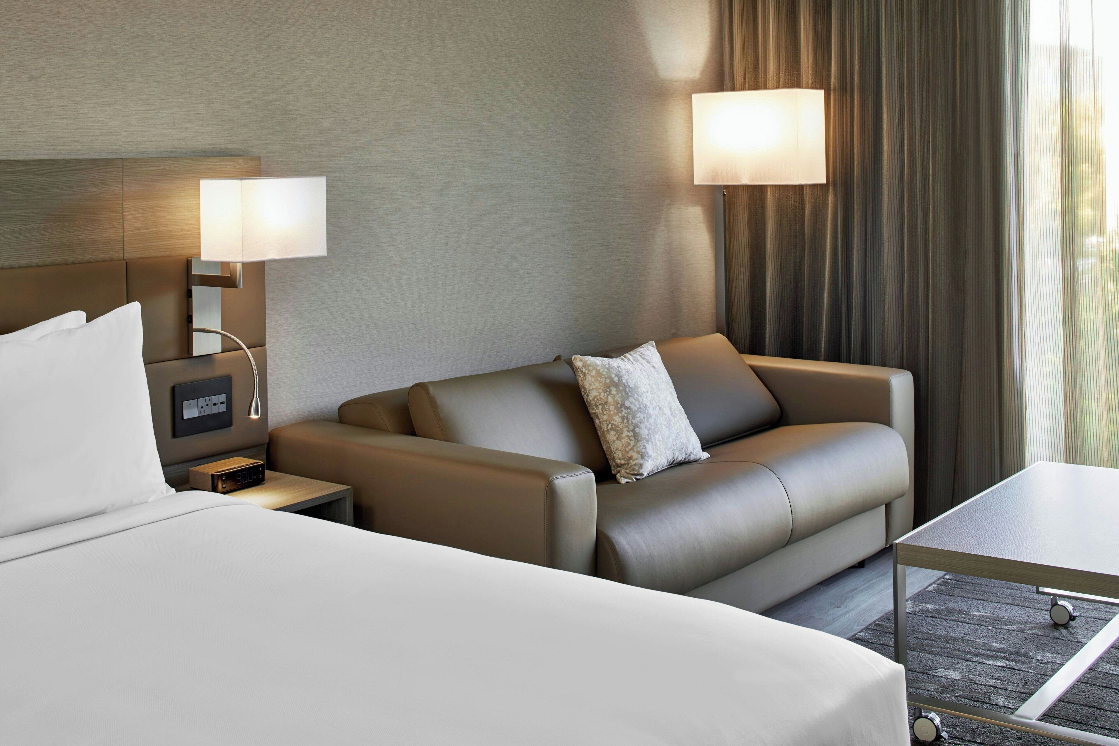 A hotel room with a bed, a sofa, a small table, a nightstand with a lamp, and a wall-mounted control panel.