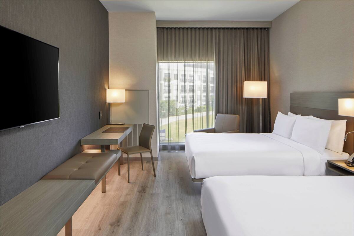 A hotel room with a TV, two beds, a desk, a chair, and a window with curtains.
