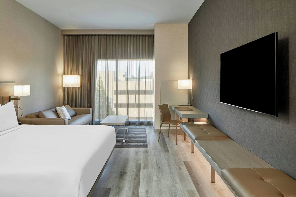A hotel room with a bed, a sofa, a desk, a chair, a TV, and a large window with curtains.