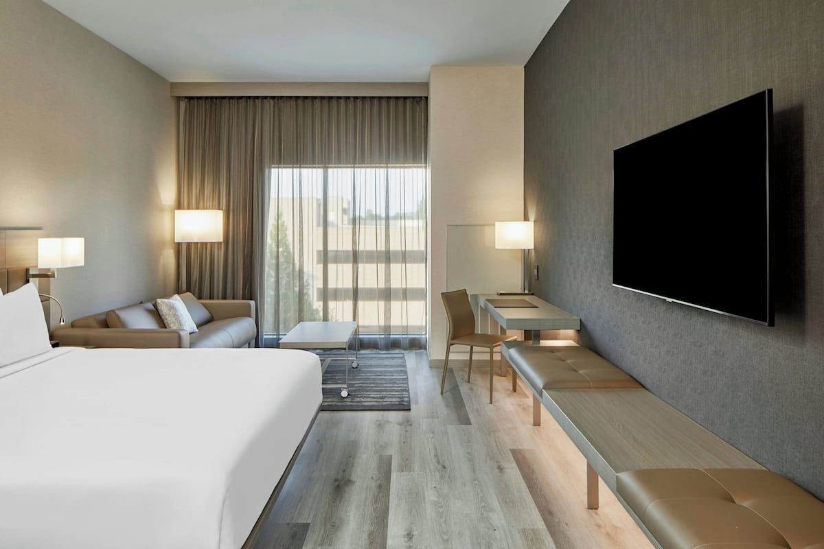 A hotel room with a bed, a sofa, a desk, a chair, a TV, and a large window with curtains.