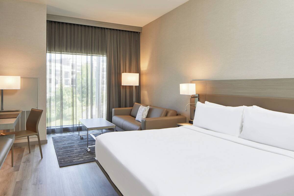 A hotel room with a large bed, a sofa, a desk, and a chair. There are two lamps on either side of the sofa.