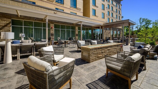 An outdoor patio with wicker furniture and a stone fireplace, surrounded by a building with multiple windows and a covered walkway. - Courtyard by Marriott Raleigh Cary Crossroads (Cary)