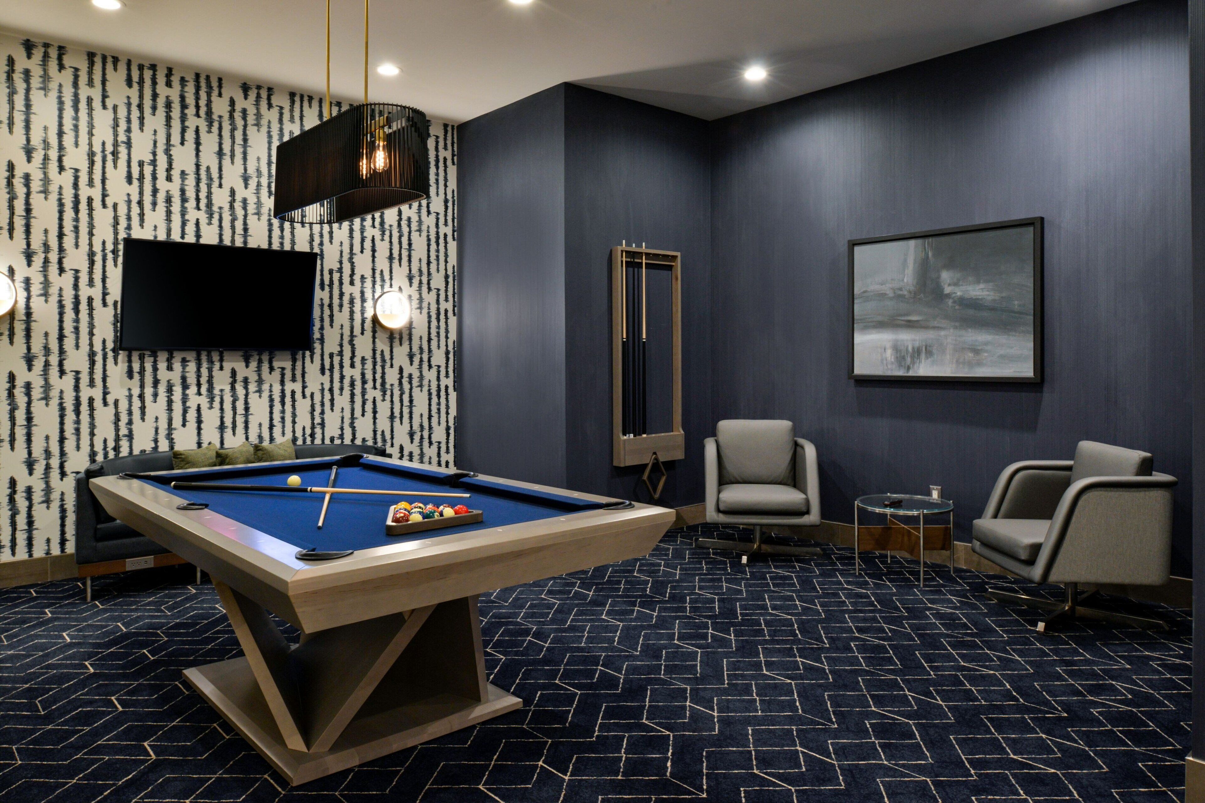 Una mesa de billar con superficie de fieltro azul, una cuesa de billar y un juego de bolas. Hay un televisor montado en la pared, un cuadro abstracto enmarcado y dos sillas. La habitación tiene una alfombra azul oscuro con un diseño geométrico.