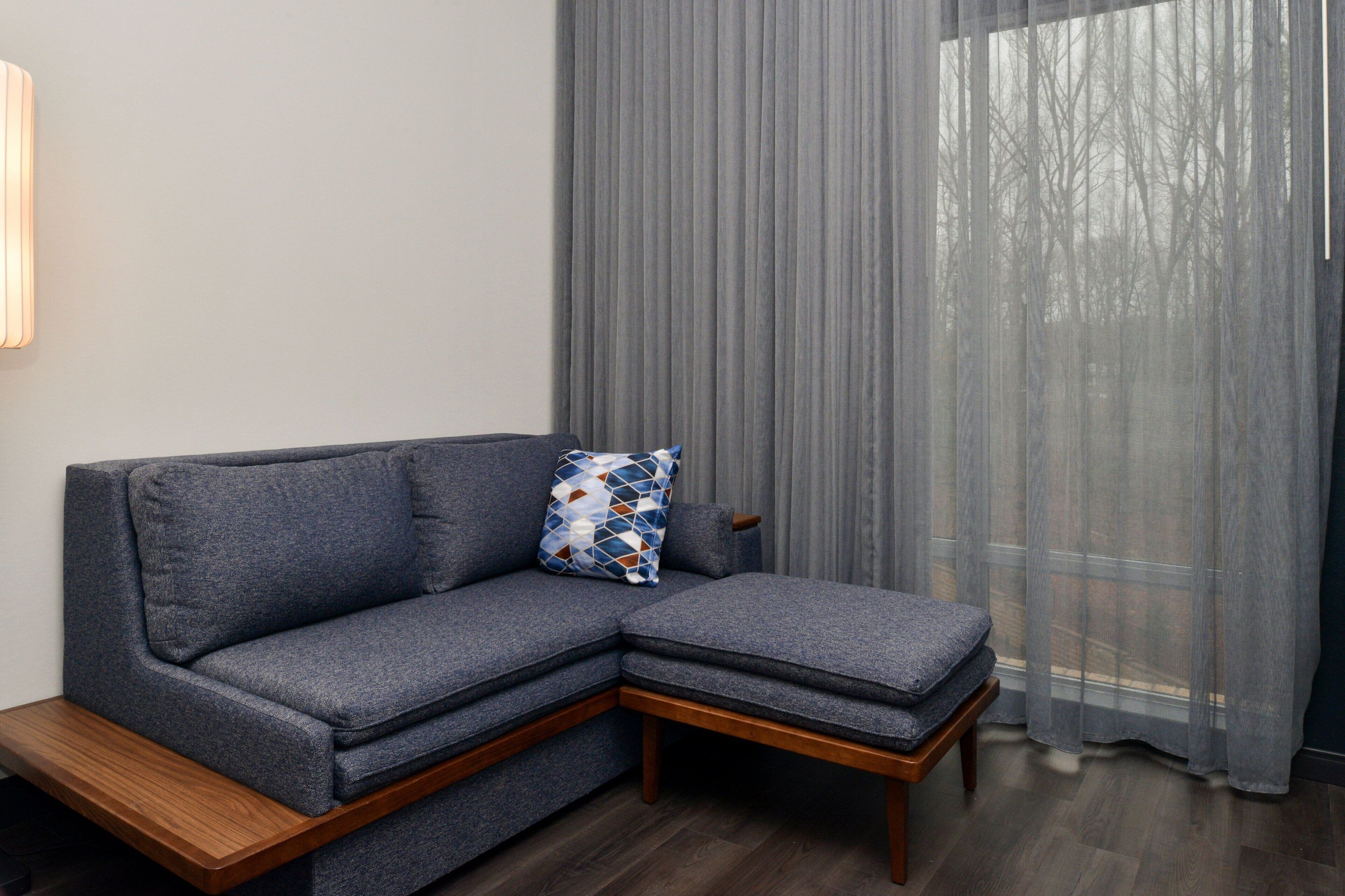 A sectional sofa with a wooden base, a patterned pillow, and a window with sheer curtains.