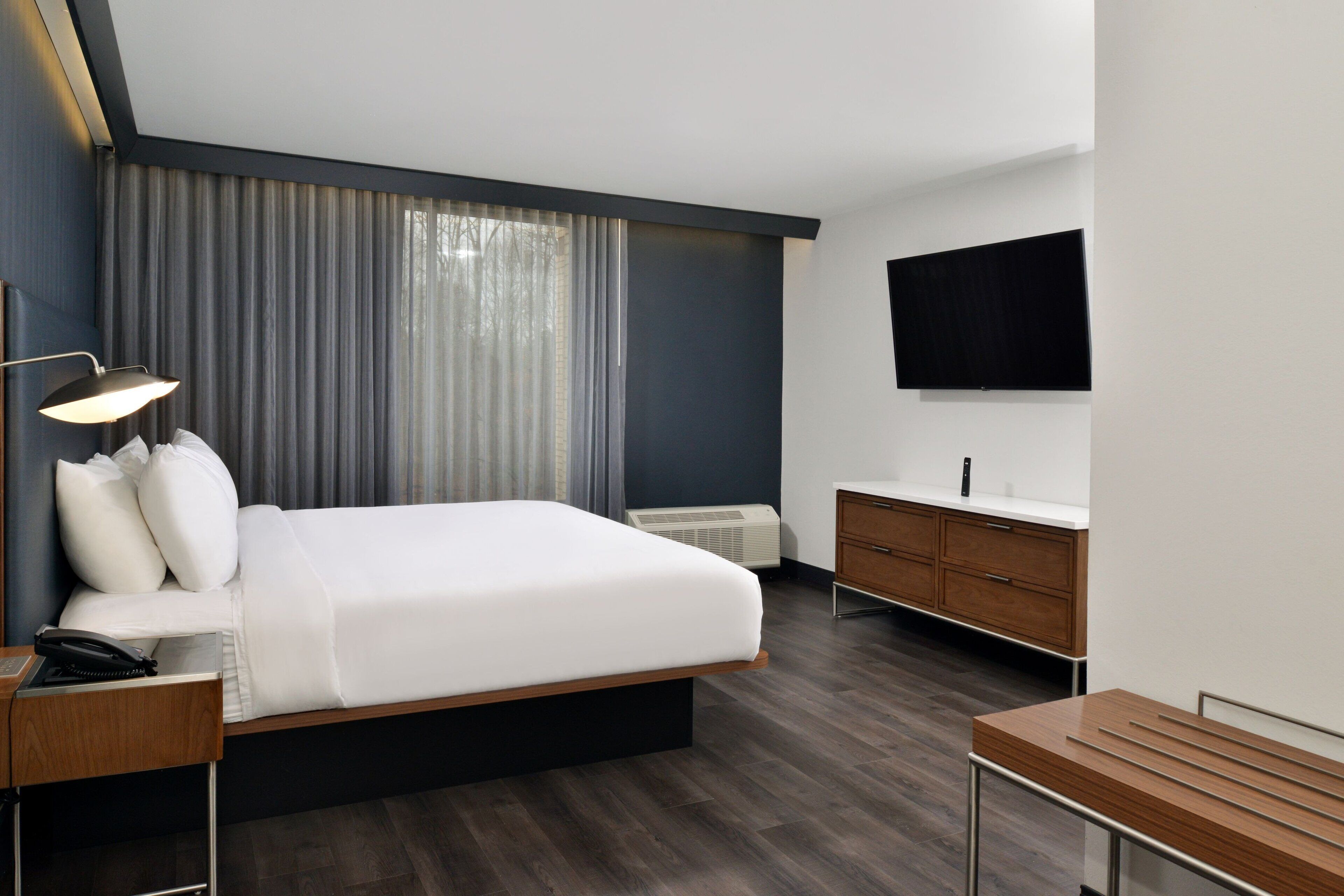 Een moderne hotelkamer met een groot bed, een nachtkastje met een lamp, een houten bankje, een badkamer met spiegel en een flatscreen televisie aan de muur.