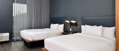 A hotel room with two beds, a large window with curtains, a bedside table with a lamp, and a wall-mounted phone.
