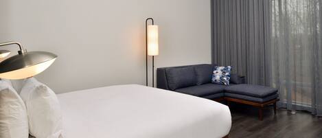 A hotel room with a bed, a desk lamp, a modern floor lamp, a grey sofa, and a large window with curtains.
