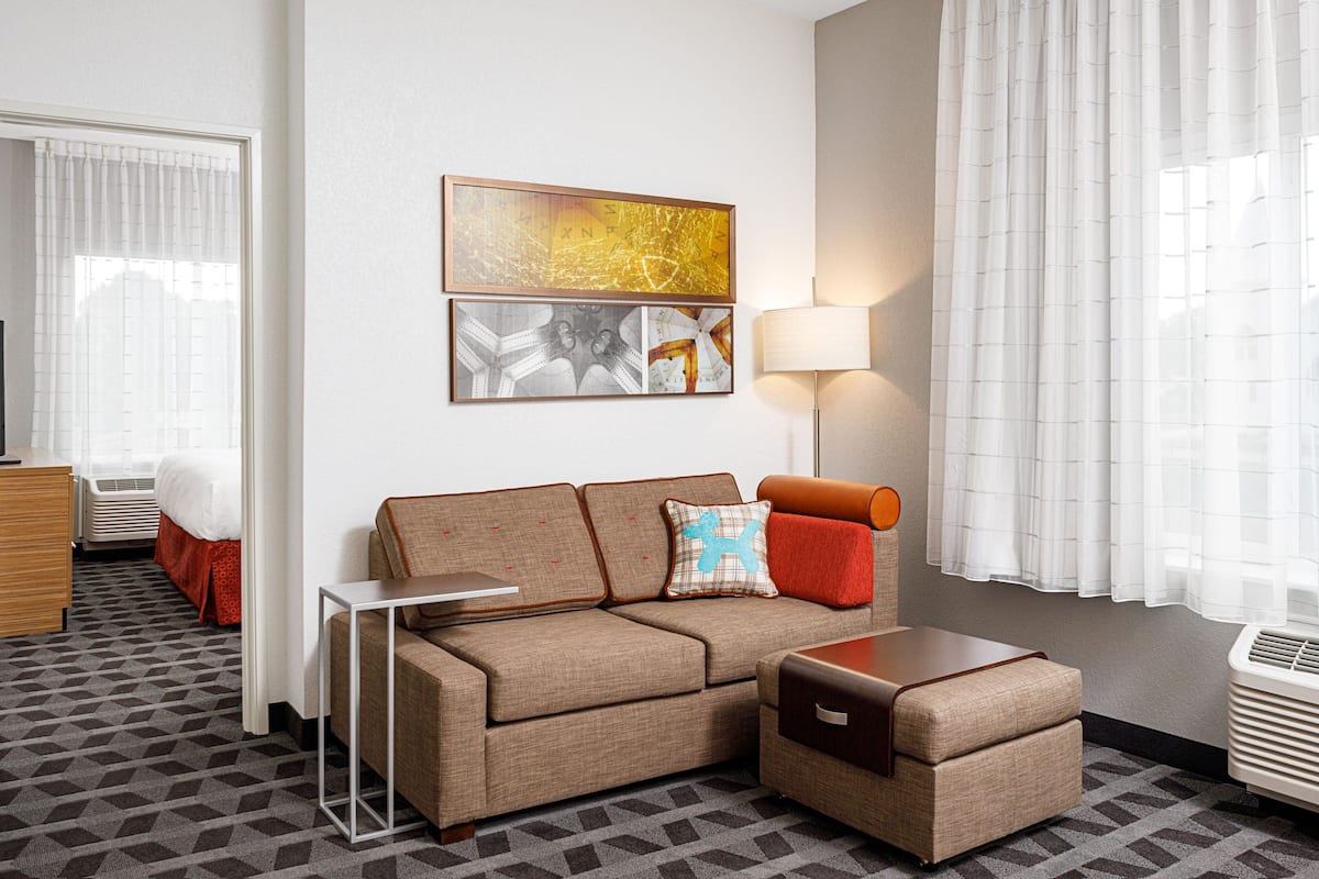 A hotel room with a sofa, a small table, a lamp, and a bed with a red accent.