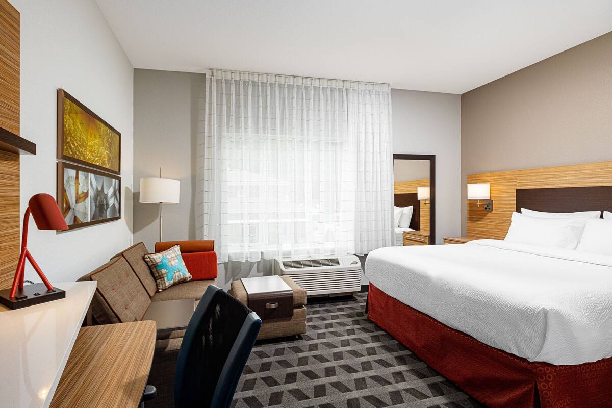 A hotel room with a bed, a desk, a chair, a small table, a lamp, a mirror, and a window with curtains.