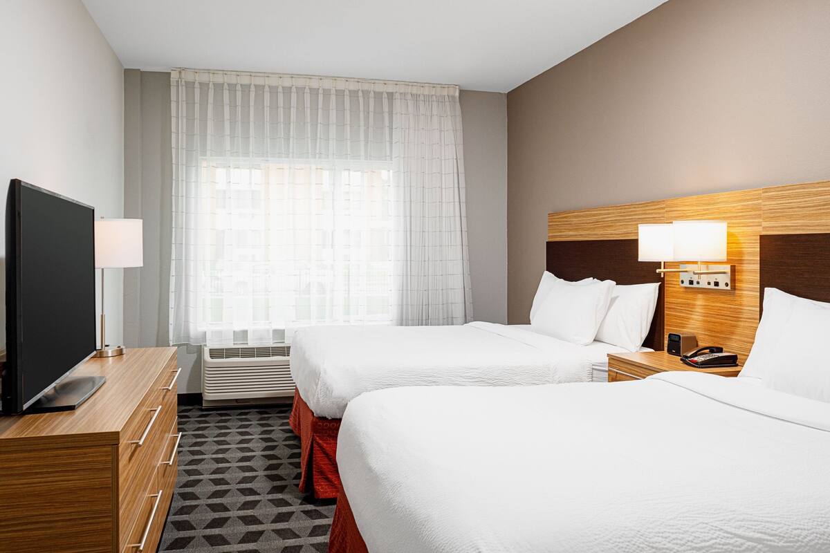 A hotel room with two beds, a TV, a bedside table with a lamp, and a wall-mounted phone.
