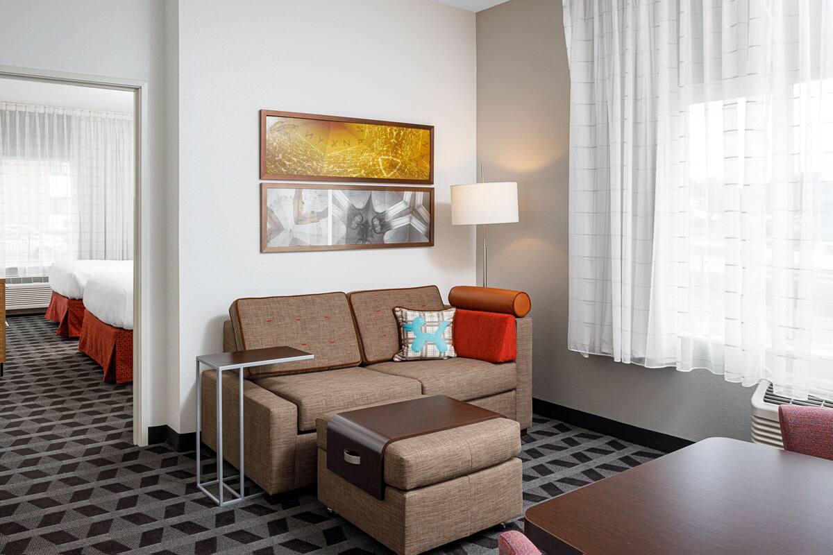 A hotel room with a sofa, a small table, a lamp, and a view of the outside through the window.