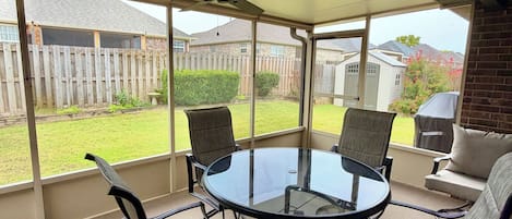 A covered patio area with a glass table and four chairs, a ceiling fan, and a view of a backyard with a wooden fence and a house.