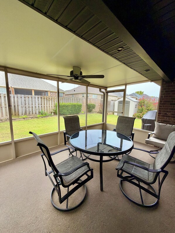 A covered patio area with a glass table and four chairs, a ceiling fan, and a view of a backyard with a wooden fence and a house.