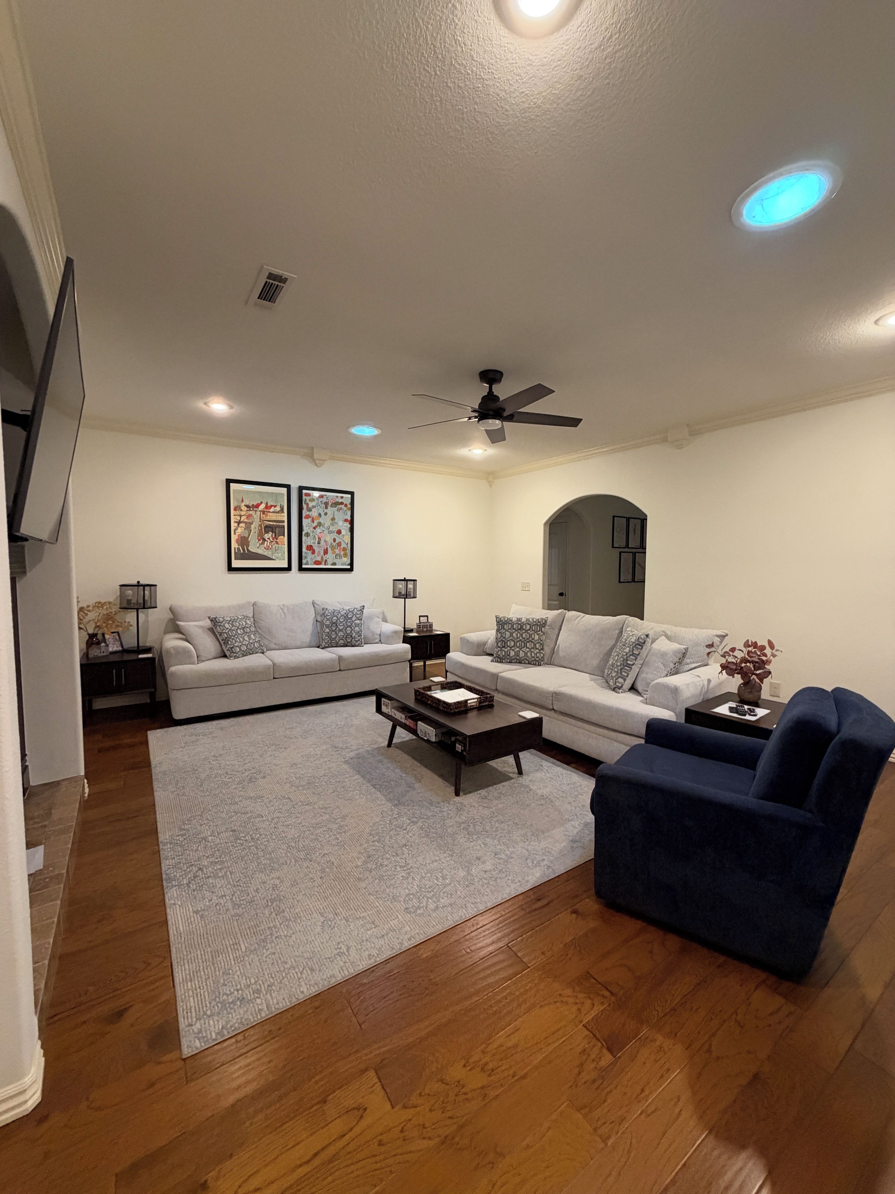 A living room with a sofa, coffee table, and blue armchair, wooden flooring, and recessed ceiling lights.