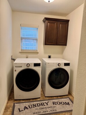 A laundry room with two white LG washing machines, a window with blinds, and a brown cabinet above.