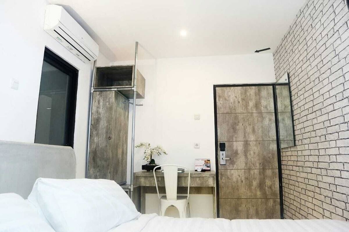 Una habitación de hotel moderna con una cama, un escritorio, una silla, un equipo de aire acondicionado, un armario y una pared de ladrillo.