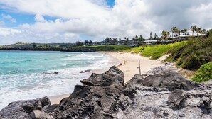 Una playa con primer plano rocoso, aguas turquesas cristalinas y una vista distante de un resort con varios edificios y palmeras.