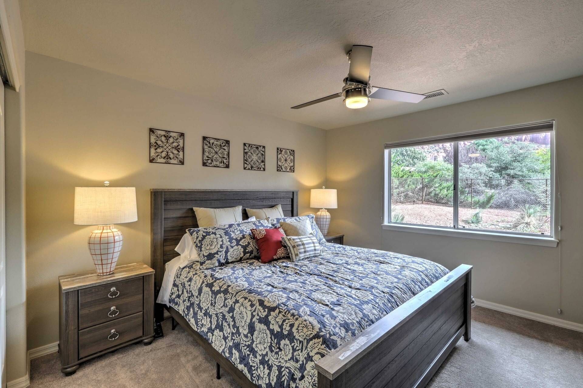 A bedroom with a large bed, two bedside tables with lamps, a ceiling fan, and a window with a view of greenery.