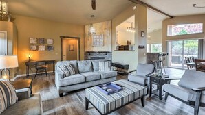 A spacious living room with a large gray sofa, a striped ottoman, and a coffee table. There is a dining area with chairs and a table, and a kitchen area with a countertop and hanging lights.