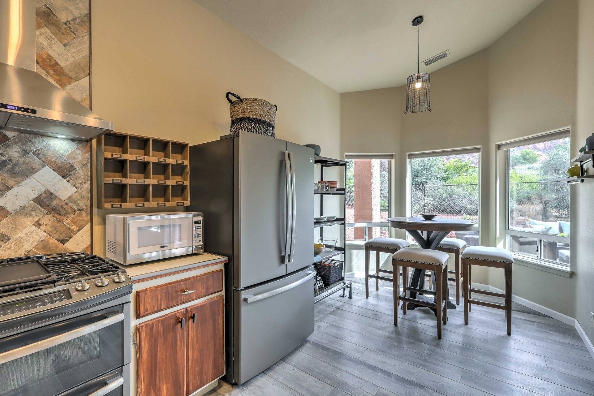 A modern kitchen with a built-in microwave, stainless steel refrigerator, and gas stove. There is a wooden cabinet with a herringbone-patterned backsplash, and a dining area with a table and chairs.