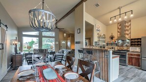 A spacious dining area with a long wooden table set for six, a kitchen with modern appliances, and a large window providing natural light.