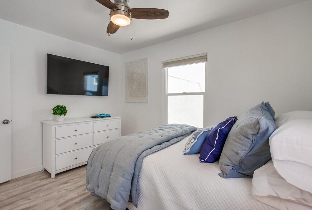 A bedroom with a bed, a white dresser, a ceiling fan, a TV, and a window.