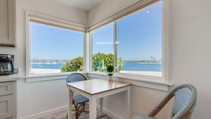 A corner room with a wooden table and two wicker chairs, large windows with a view of the sea and greenery, and a white cabinet with a coffee maker.